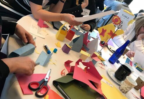Participants work together on a creative task involving colorful paper cutouts during the Helping Hands team building event. They collaborate and discuss their progress while assembling materials for care kits.