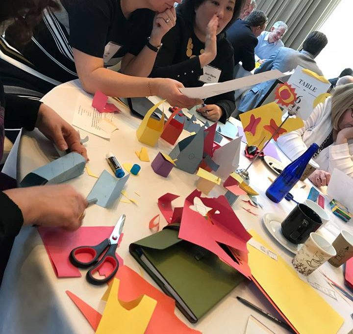 Participants work together on a creative task involving colorful paper cutouts during the Helping Hands team building event. They collaborate and discuss their progress while assembling materials for care kits.