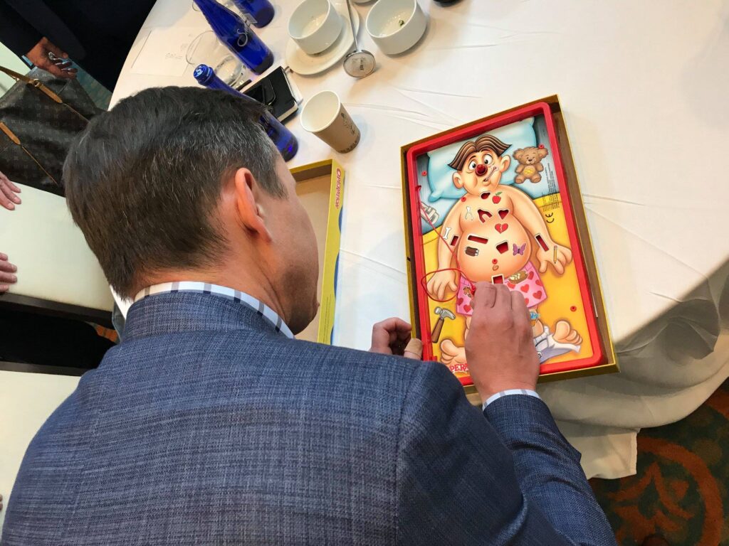 A participant plays the game 'Operation' at a table during the Helping Hands team building event. The game is part of a fun challenge to earn supplies for assembling personal care kits for those in need.