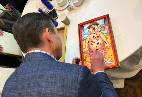 A participant plays the game 'Operation' at a table during the Helping Hands team building event. The game is part of a fun challenge to earn supplies for assembling personal care kits for those in need.