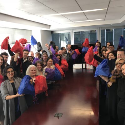 A group of participants stands around a long table, proudly holding up colorful drawstring bags filled with care kits during the Helping Hands team building event. They are smiling and celebrating their effort to support the community. thumbnail
