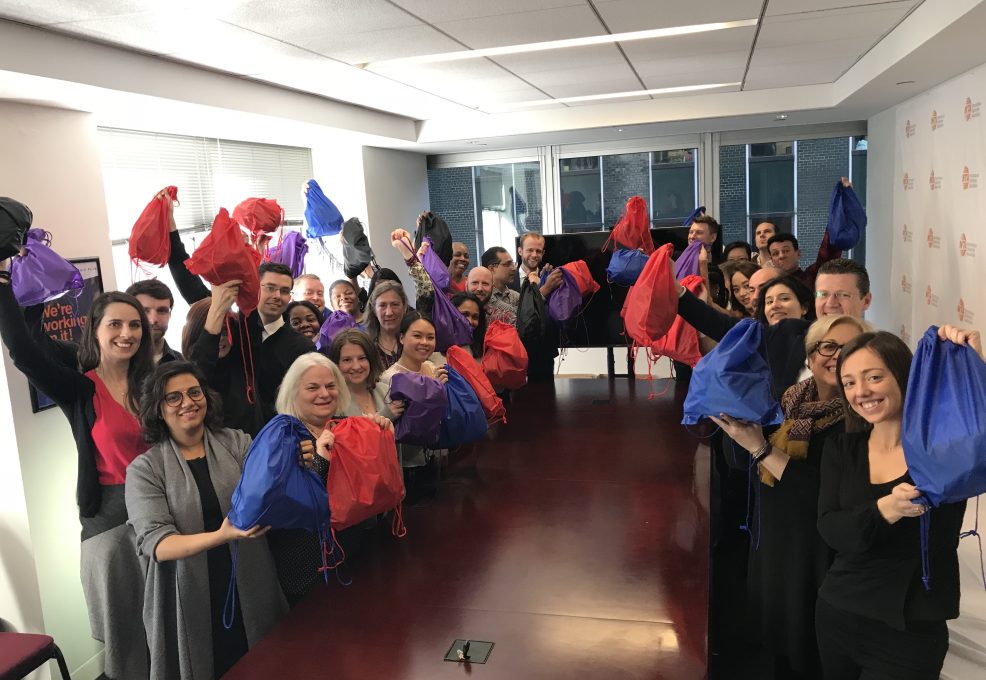 A group of participants stands around a long table, proudly holding up colorful drawstring bags filled with care kits during the Helping Hands team building event. They are smiling and celebrating their effort to support the community.