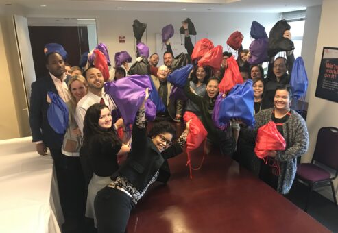 A large group of participants proudly holds up colorful drawstring bags filled with care kits they assembled during the Helping Hands team building event. The group is smiling and celebrating their contributions to the community.