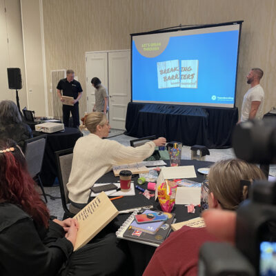 Participants sit at tables holding wooden boards while watching a facilitator prepare for the board-breaking demonstration during a team building event focused on breaking through barriers. thumbnail
