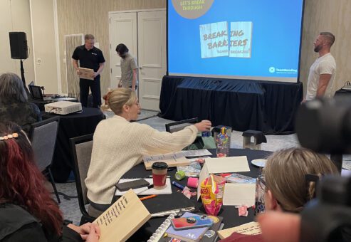 Participants sit at tables holding wooden boards while watching a facilitator prepare for the board-breaking demonstration during a team building event focused on breaking through barriers.