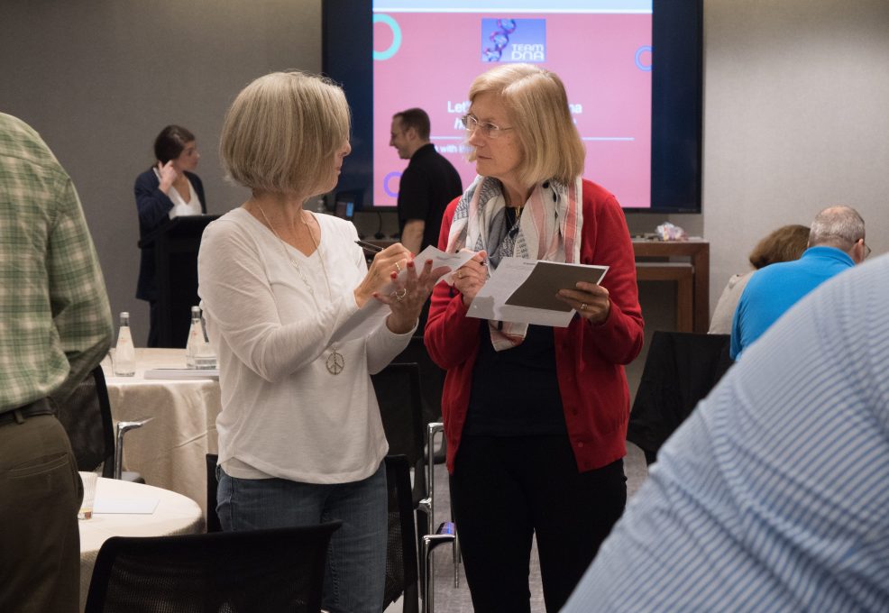 Two participants discussing and exchanging ideas during a Team DNA or Leadership DNA session, with other attendees engaged in the background.