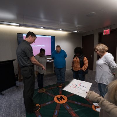Participants engaging in a Team DNA activity, working together to solve a challenge while a facilitator leads the session with a visual display in the background. thumbnail