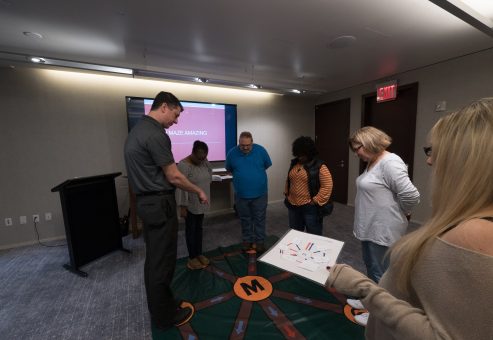 Participants engaging in a Team DNA activity, working together to solve a challenge while a facilitator leads the session with a visual display in the background.