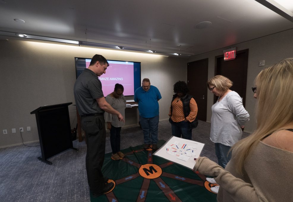 Participants engaging in a Team DNA activity, working together to solve a challenge while a facilitator leads the session with a visual display in the background.
