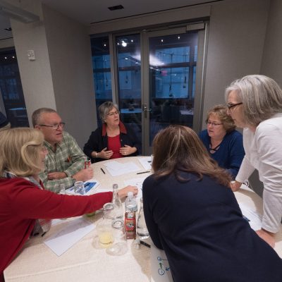 A group of participants collaborating at a table during a Team DNA workshop, discussing and working on materials with a facilitator nearby. thumbnail