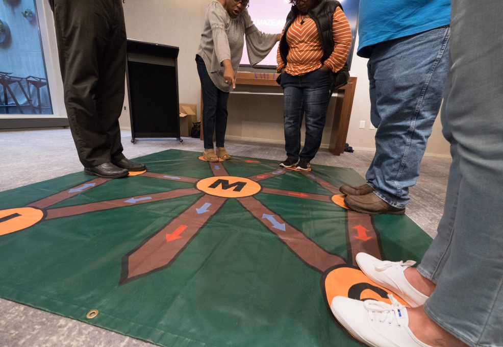 Participants engaging in an interactive Team and Leadership DNA activity, using a large, colorful floor mat designed to explore key team dynamics and enhance communication.