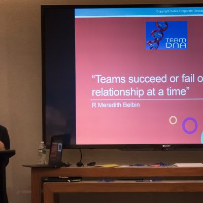 A facilitator leading a Team DNA session with a presentation slide on the screen that reads: 'Teams succeed or fail one relationship at a time' - R Meredith Belbin. thumbnail
