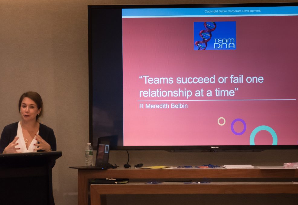 A facilitator leading a Team DNA session with a presentation slide on the screen that reads: 'Teams succeed or fail one relationship at a time' - R Meredith Belbin.