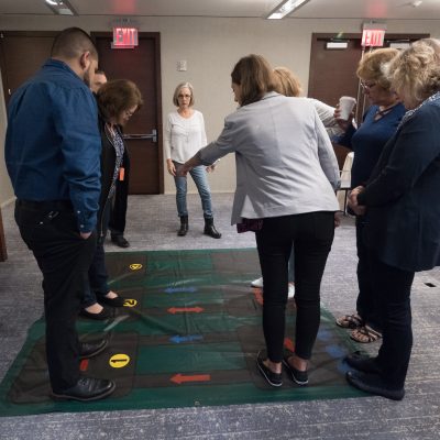 A group participating in a Team DNA activity, standing on a large interactive mat with color-coded arrows, while a facilitator provides instructions. thumbnail