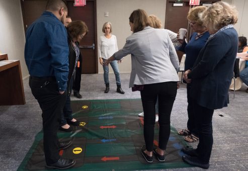 A group participating in a Team DNA activity, standing on a large interactive mat with color-coded arrows, while a facilitator provides instructions.