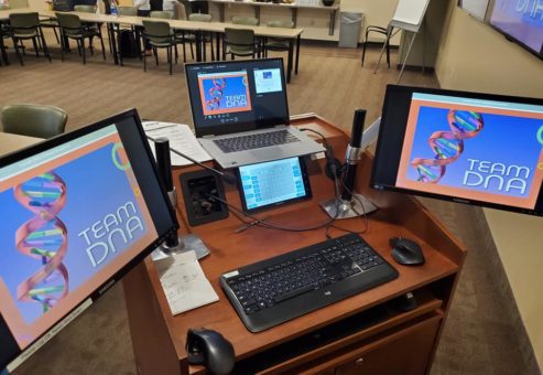 A workstation set up for the Team DNA session, featuring multiple screens displaying the event's branding and presentation materials.