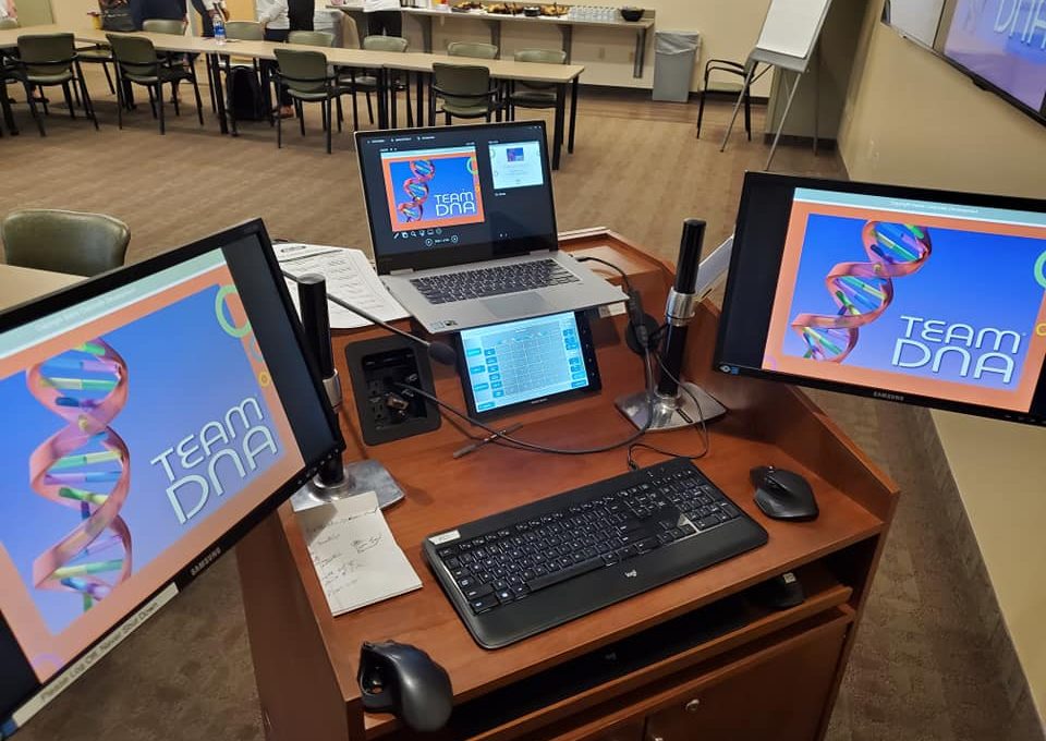 A workstation set up for the Team DNA session, featuring multiple screens displaying the event's branding and presentation materials.