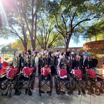 A large group of participants stands outdoors behind a row of assembled wheelchairs decorated with patriotic red, white, and blue ribbons during the Wheelchairs In Motion team building event. The group is celebrating their successful charitable effort. thumbnail
