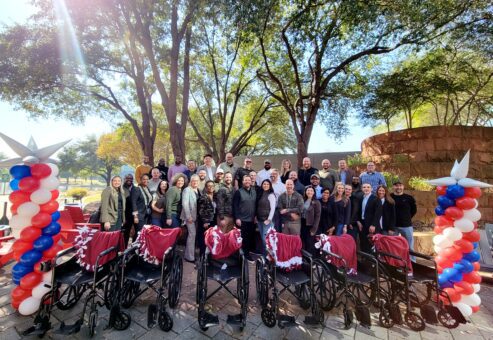 A large group of participants stands outdoors behind a row of assembled wheelchairs decorated with patriotic red, white, and blue ribbons during the Wheelchairs In Motion team building event. The group is celebrating their successful charitable effort.