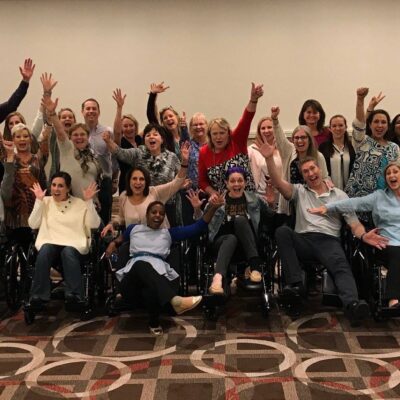 A large group of participants enthusiastically raises their hands while sitting in wheelchairs, celebrating the completion of their Wheelchairs In Motion team building event. The group is smiling and excited after successfully assembling the wheelchairs for charity. thumbnail