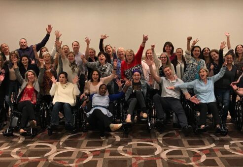 A large group of participants enthusiastically raises their hands while sitting in wheelchairs, celebrating the completion of their Wheelchairs In Motion team building event. The group is smiling and excited after successfully assembling the wheelchairs for charity.