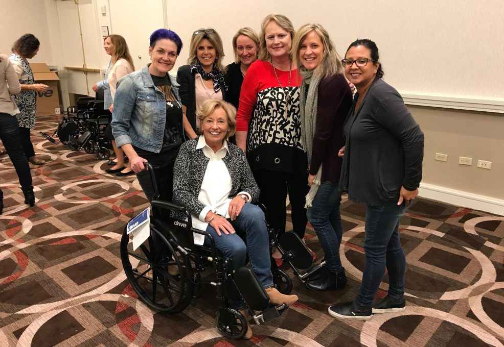 A group of participants smiles and gathers around a decorated wheelchair during the Wheelchairs In Motion team building event. One person sits in the wheelchair as the others pose together, celebrating their successful charitable effort.
