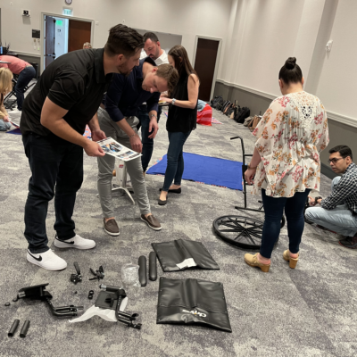 Participants gather around wheelchair parts, working together to assemble them during the Wheelchairs In Motion team building event. The room is filled with groups collaborating on similar tasks as part of the charitable activity. thumbnail