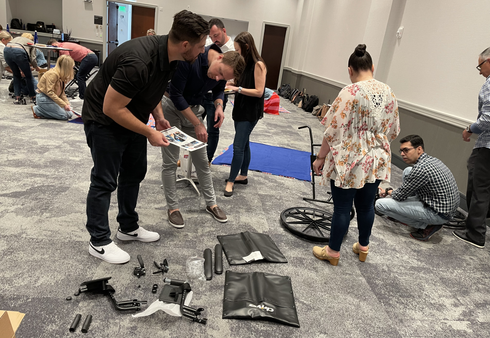 Participants gather around wheelchair parts, working together to assemble them during the Wheelchairs In Motion team building event. The room is filled with groups collaborating on similar tasks as part of the charitable activity.