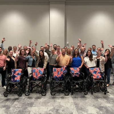 A large group of participants stands behind a row of assembled wheelchairs decorated with American flag blankets, celebrating their achievement during the Wheelchairs In Motion team building event. The group is smiling and raising their hands in celebration. thumbnail