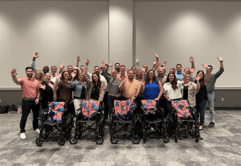 A large group of participants stands behind a row of assembled wheelchairs decorated with American flag blankets, celebrating their achievement during the Wheelchairs In Motion team building event. The group is smiling and raising their hands in celebration.