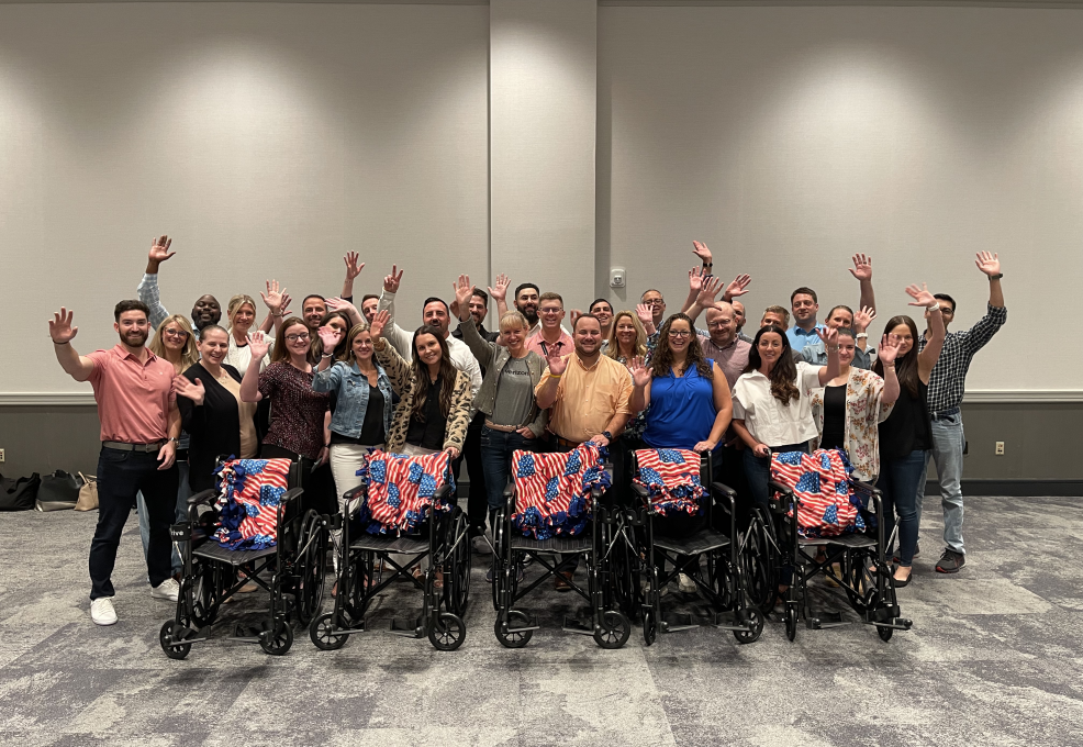 A large group of participants stands behind a row of assembled wheelchairs decorated with American flag blankets, celebrating their achievement during the Wheelchairs In Motion team building event. The group is smiling and raising their hands in celebration.