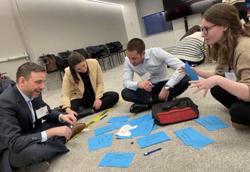 A group of participants sits on the floor, engaged in a collaborative discussion and organizing blue cards during a Wheelchairs In Motion team building event. They are brainstorming and working through tasks as part of the charitable activity.