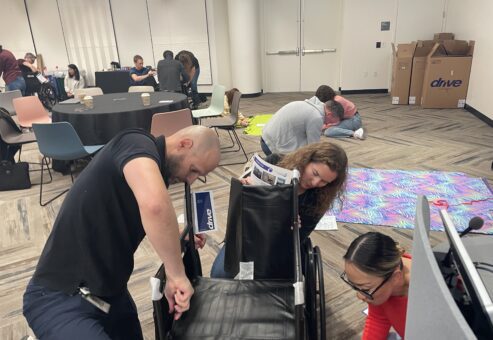 A group of participants collaborates to assemble a wheelchair during the Wheelchairs In Motion team building event. The activity is part of a charitable effort to donate wheelchairs to veterans and those in need.