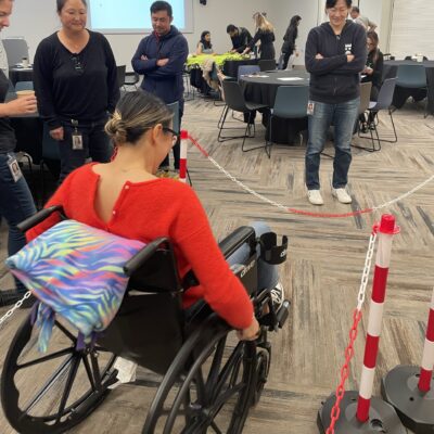 A participant navigates an obstacle course in a wheelchair during the Wheelchairs In Motion team building event. Other participants watch and support as she tests the newly assembled wheelchair. thumbnail
