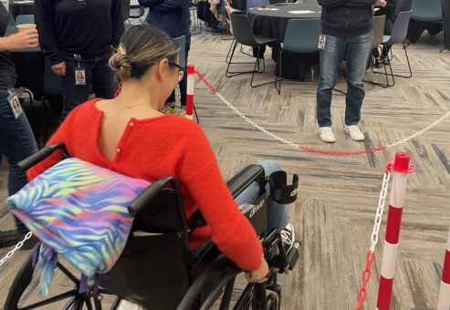 A participant navigates an obstacle course in a wheelchair during the Wheelchairs In Motion team building event. Other participants watch and support as she tests the newly assembled wheelchair.