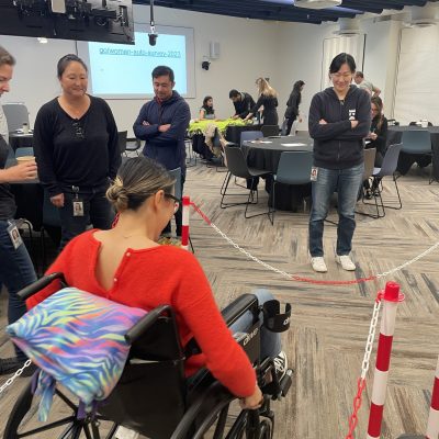 A participant navigates an obstacle course in a wheelchair during the Wheelchairs In Motion team building event. Other participants watch and support as she tests the newly assembled wheelchair. thumbnail
