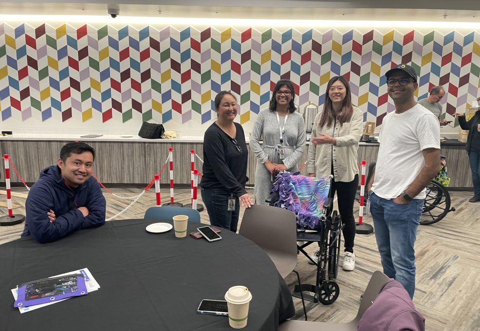 A group of participants gathers around a table in a colorful indoor space during the Wheelchairs In Motion team building event. A decorated wheelchair is nearby, and the team is smiling, enjoying the charitable activity.
