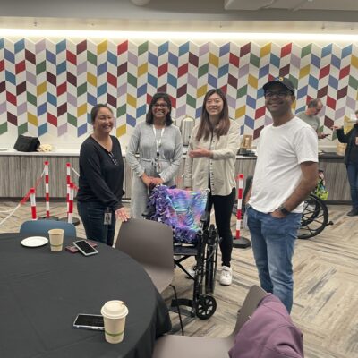 A group of participants gathers around a table in a colorful indoor space during the Wheelchairs In Motion team building event. A decorated wheelchair is nearby, and the team is smiling, enjoying the charitable activity. thumbnail
