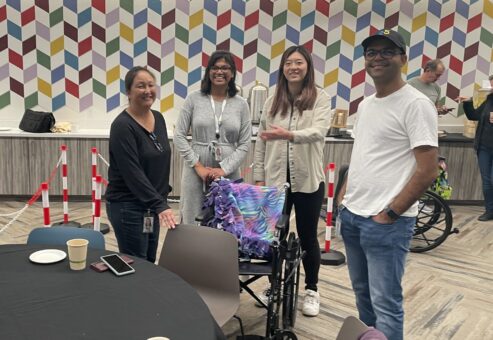 A group of participants gathers around a table in a colorful indoor space during the Wheelchairs In Motion team building event. A decorated wheelchair is nearby, and the team is smiling, enjoying the charitable activity.