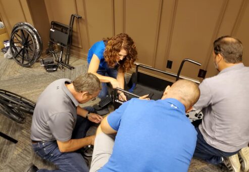 A group of participants collaborates on assembling a wheelchair during the Wheelchairs In Motion team building event. They work closely together, focusing on attaching parts to the wheelchair frame.