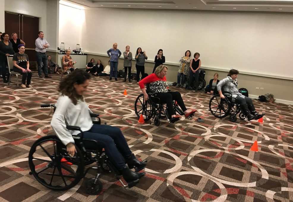 Participants race in wheelchairs through an obstacle course inside a conference room during the Wheelchairs In Motion team building event. Other attendees cheer them on as they navigate through the course.