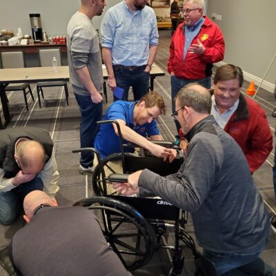 A group of participants works together to assemble a wheelchair during the Wheelchairs In Motion team building event. Several people collaborate, focusing on the task at hand, while others observe and converse in the background. thumbnail