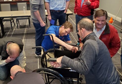 A group of participants works together to assemble a wheelchair during the Wheelchairs In Motion team building event. Several people collaborate, focusing on the task at hand, while others observe and converse in the background.