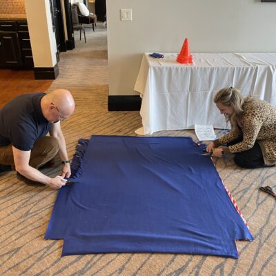 Two participants work together on the floor, cutting and preparing a large fabric piece during the Wheelchairs In Motion team building event. The fabric is part of a blanket for the wheelchair donation. thumbnail