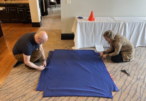 Two participants work together on the floor, cutting and preparing a large fabric piece during the Wheelchairs In Motion team building event. The fabric is part of a blanket for the wheelchair donation.