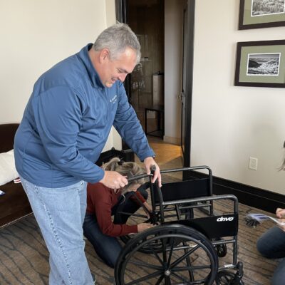 A group of participants works together to assemble a wheelchair during the Wheelchairs In Motion team building event. One participant is focused on adjusting the wheelchair, while others assist and read instructions nearby. thumbnail