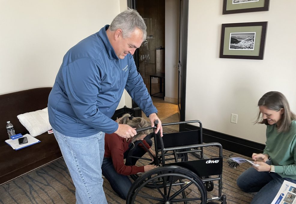 A group of participants works together to assemble a wheelchair during the Wheelchairs In Motion team building event. One participant is focused on adjusting the wheelchair, while others assist and read instructions nearby.