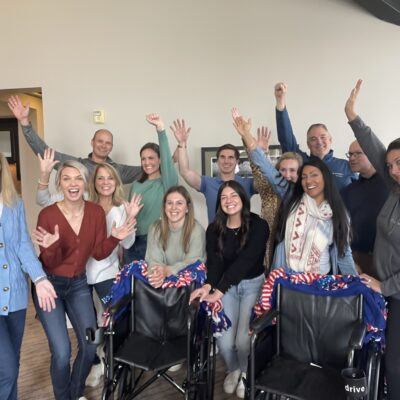 A group of participants enthusiastically raises their hands, celebrating their successful efforts during the Wheelchairs In Motion team building event. Two decorated wheelchairs are in front of the group, ready for donation. thumbnail
