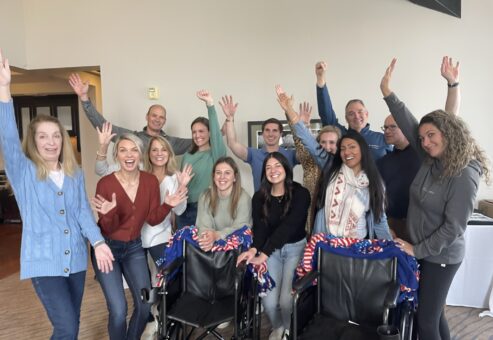 A group of participants enthusiastically raises their hands, celebrating their successful efforts during the Wheelchairs In Motion team building event. Two decorated wheelchairs are in front of the group, ready for donation.