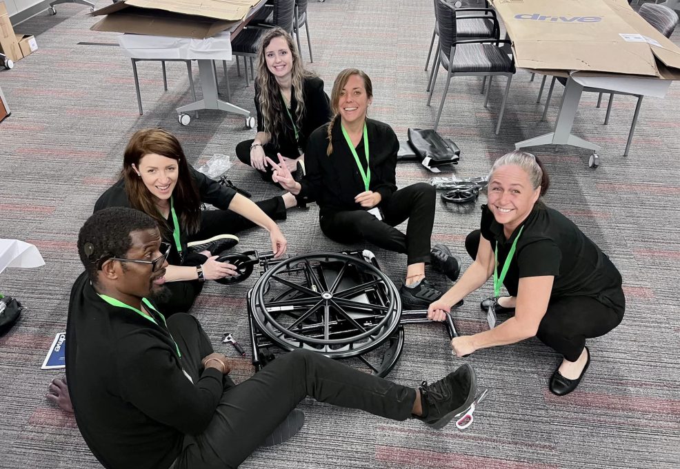 A group of smiling participants sits on the floor working together to assemble a wheelchair during the Wheelchairs In Motion team building event. The activity is part of a charitable initiative to donate wheelchairs to those in need.